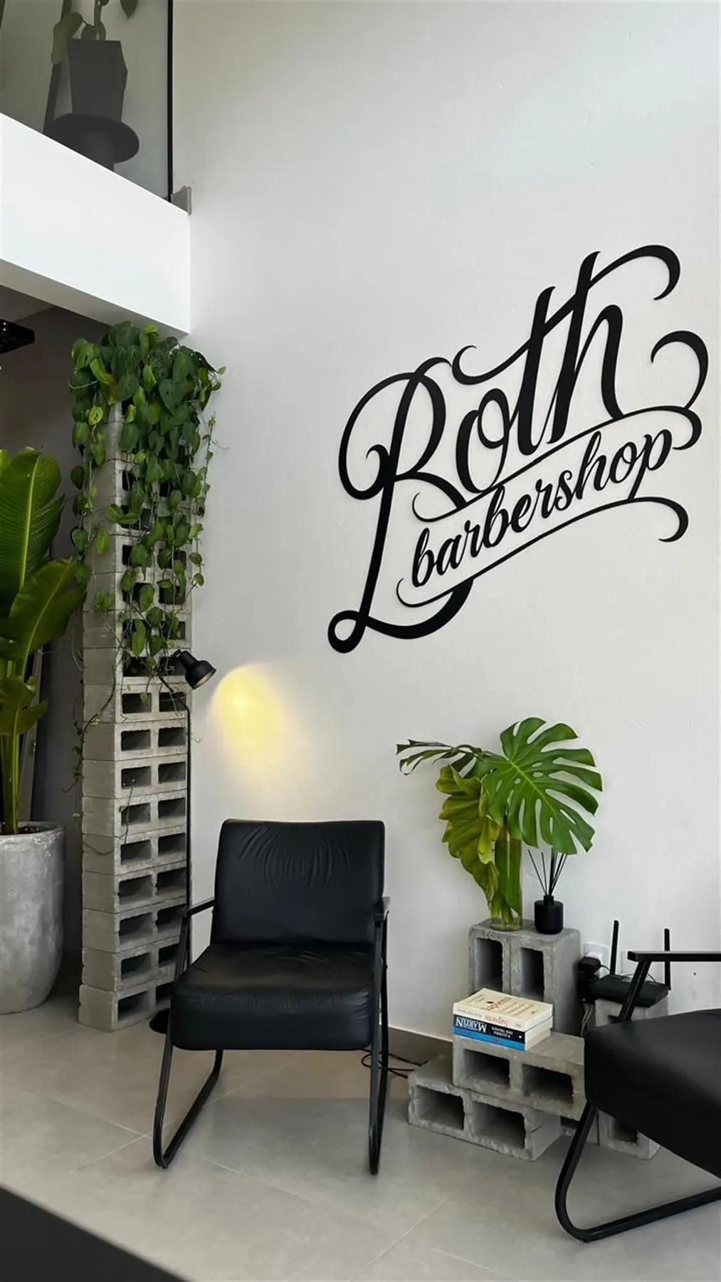 Parede assinada Both Barbershop, com poltronas de espera em couro preto e pilar de blocos de cimento com plantas